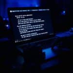 Young musician bathed in blue light using an AI lyrics generator on their laptop, with songwriting structures on screen and handwritten notes nearby, surrounded by shadowy instruments.